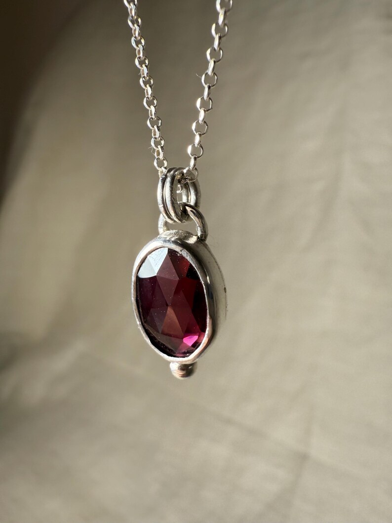 May include: A silver necklace featuring a pendant with a faceted, oval-shaped, deep red gemstone. The pendant is set in a silver bezel and hangs from a delicate silver chain. The gemstone has a rich, dark color, and the necklace is displayed against a neutral background.