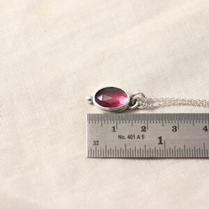 May include: A silver pendant necklace with a faceted, oval-shaped, deep pink gemstone. The pendant is approximately 0.75 inches long. The necklace is on a light beige background.