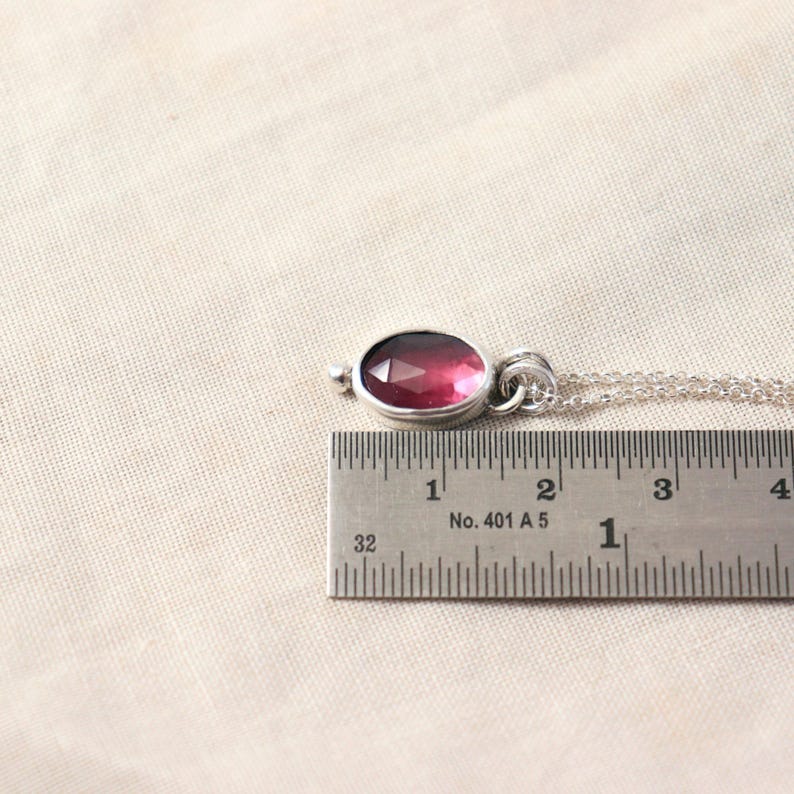 May include: A silver pendant necklace with a faceted, oval-shaped, deep pink gemstone. The pendant is approximately 0.75 inches long. The necklace is on a light beige background.