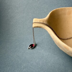 May include: A silver necklace with a small, oval-shaped pendant featuring a deep red gemstone. The pendant is suspended from a delicate chain. The necklace rests on a light brown, curved surface against a blue background.