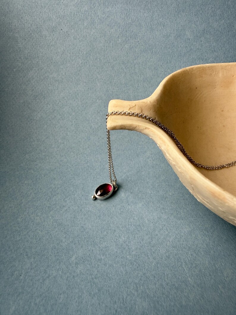 May include: A silver necklace with a small, oval-shaped pendant featuring a deep red gemstone. The pendant is suspended from a delicate chain. The necklace rests on a light brown, curved surface against a blue background.