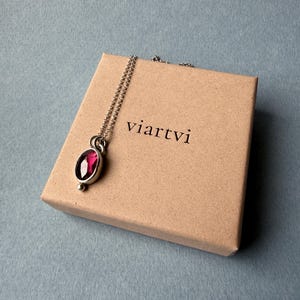 May include: A silver pendant necklace with a large, oval, ruby-colored gemstone. The necklace rests on a small, tan cardboard box with the word "viartvi" printed on it. The background is a muted blue-gray.