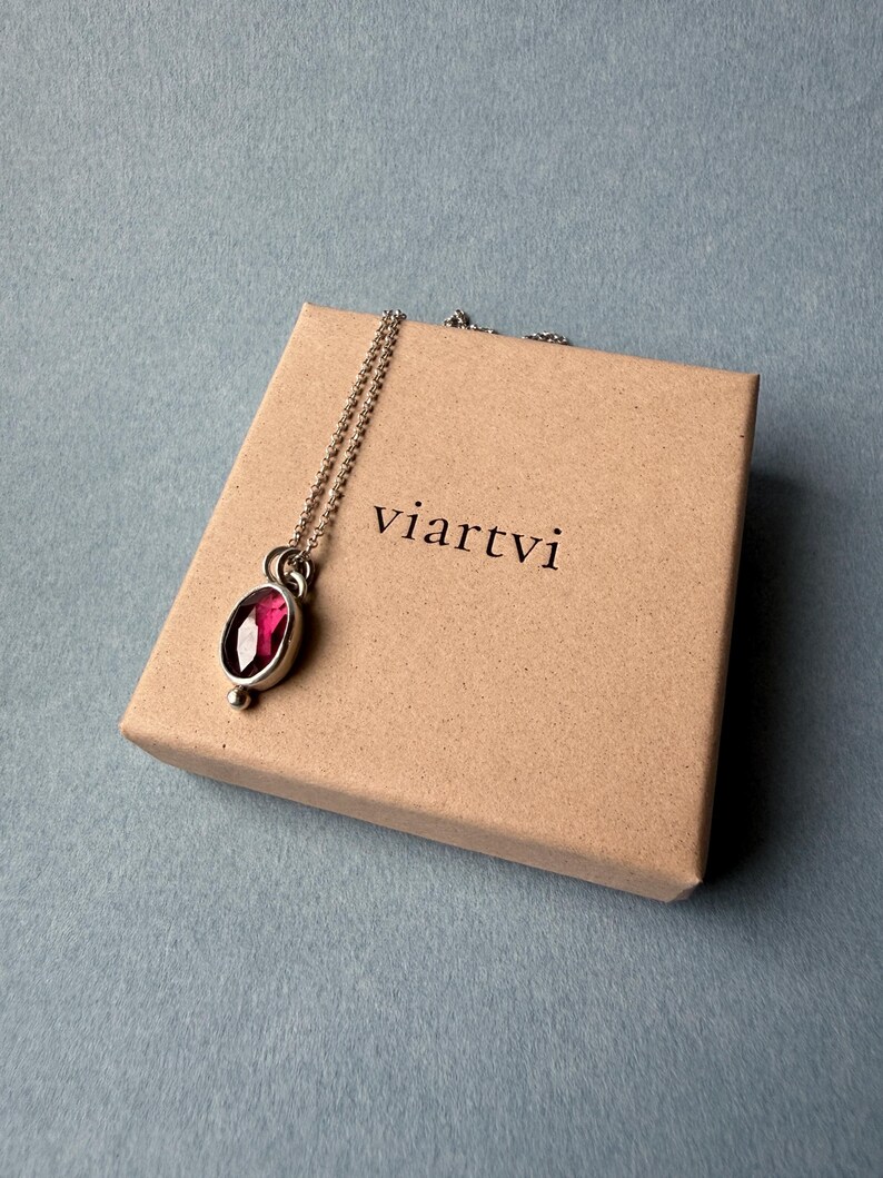 May include: A silver pendant necklace with a large, oval, ruby-colored gemstone. The necklace rests on a small, tan cardboard box with the word "viartvi" printed on it. The background is a muted blue-gray.