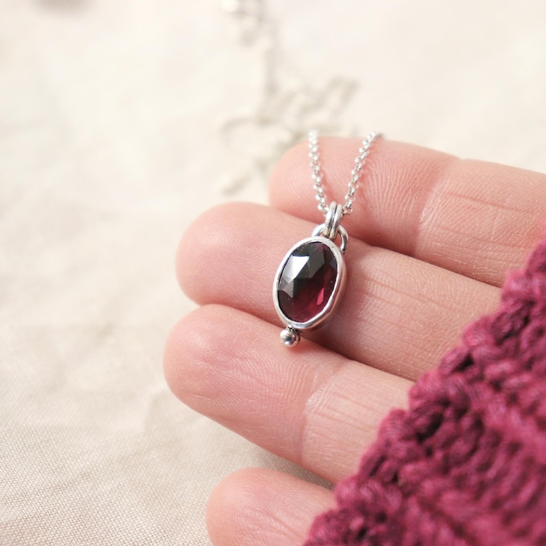 May include: A silver necklace with a dark red oval gemstone pendant. The pendant is set in a silver bezel and has a small silver bead at the bottom. The necklace is on a delicate silver chain.