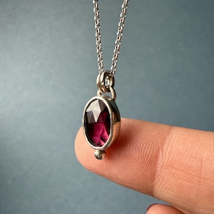 May include: A silver necklace with a faceted, oval-shaped, deep red gemstone pendant. The pendant is set in a silver bezel and hangs from a delicate silver chain. A small silver bead is at the bottom of the pendant.