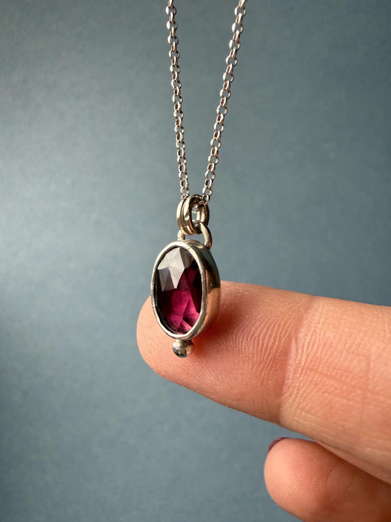 May include: A silver necklace with a faceted, oval-shaped, deep red gemstone pendant. The pendant is set in a silver bezel and hangs from a delicate silver chain. A small silver bead is at the bottom of the pendant.