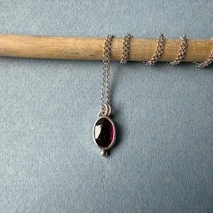 May include: A silver necklace with a small, oval, deep red gemstone pendant. The pendant is set in a silver bezel and has a small silver bead at the bottom. The chain is delicate and silver.