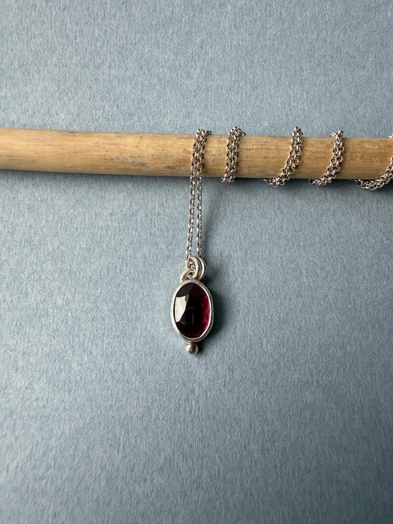 May include: A silver necklace with a small, oval, deep red gemstone pendant. The pendant is set in a silver bezel and has a small silver bead at the bottom. The chain is delicate and silver.