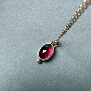 May include: A silver necklace with a pendant featuring a deep red, oval-shaped gemstone. The gemstone is set in a silver bezel and has a small silver bead at the bottom. The chain is delicate and silver.