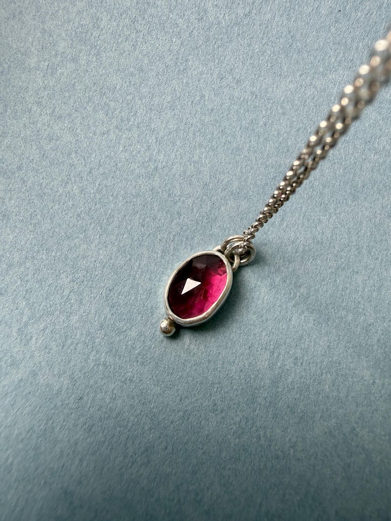 May include: A silver necklace with a pendant featuring a deep red, oval-shaped gemstone. The gemstone is set in a silver bezel and has a small silver bead at the bottom. The chain is delicate and silver.