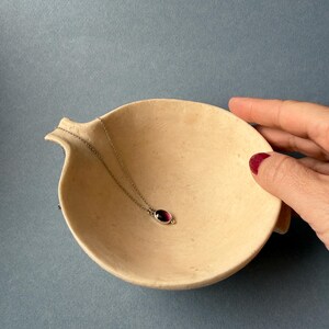 May include: A silver necklace with a small, oval, dark red gemstone pendant rests in a beige, shallow, ceramic bowl. The bowl has a curved shape with a small spout on one side. The background is a solid, muted blue.