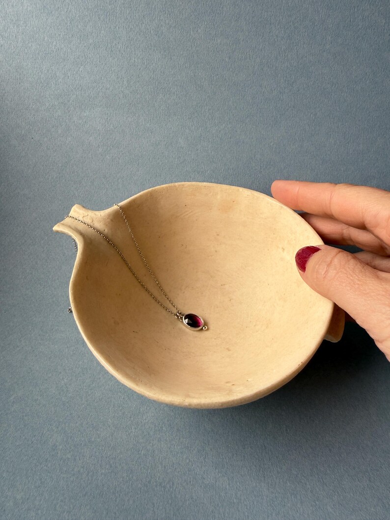 May include: A silver necklace with a small, oval, dark red gemstone pendant rests in a beige, shallow, ceramic bowl. The bowl has a curved shape with a small spout on one side. The background is a solid, muted blue.