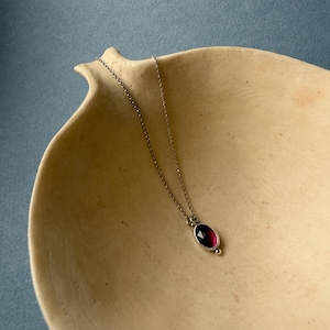 May include: A silver necklace with a deep red oval gemstone pendant. The pendant is set in a silver bezel and hangs from a delicate chain. The necklace is displayed on a beige, organic-shaped dish against a blue background.