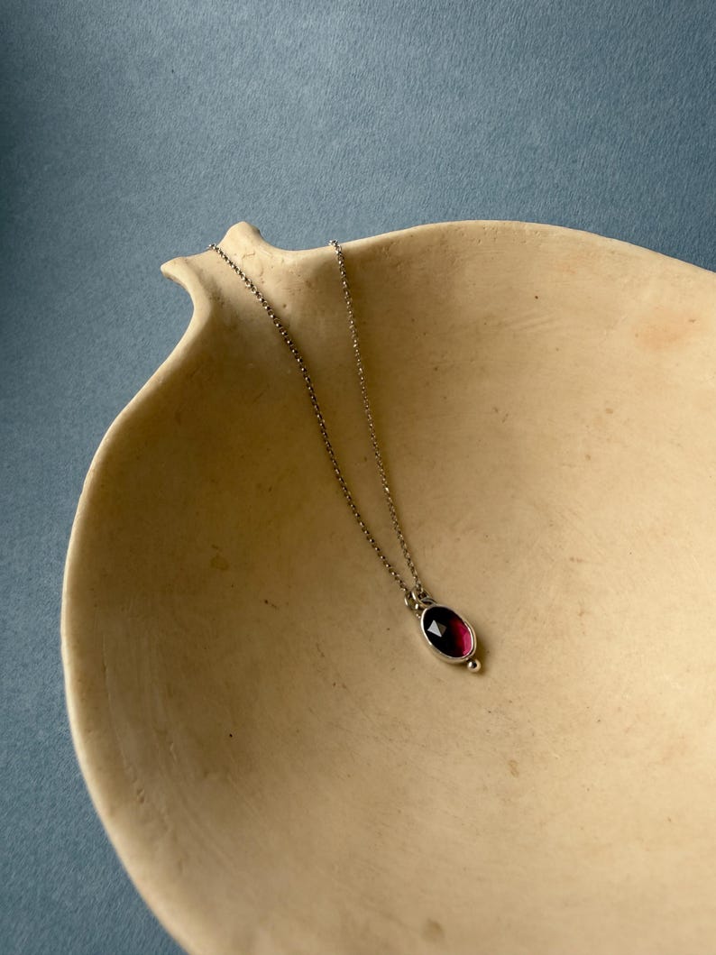 May include: A silver necklace with a deep red oval gemstone pendant. The pendant is set in a silver bezel and hangs from a delicate chain. The necklace is displayed on a beige, organic-shaped dish against a blue background.