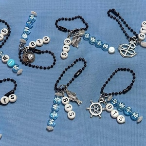 May include: Seven black ball chain keychains featuring silver-toned nautical charms like anchors, starfish, and seashells. Each keychain includes white and light blue letter beads spelling out BTS and SWIM, arranged in various configurations.