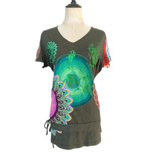 May include: An olive green V-neck t-shirt featuring a large central graphic of concentric green circles, surrounded by colorful floral and mandala-inspired patterns in shades of pink, teal, and orange, with a drawstring detail at the hem.
