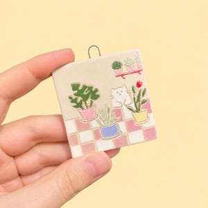 Op de afbeelding: Een vierkant keramieken ornament met een witte kat die tussen potplanten op een roze en wit geblokte ondergrond zit. Het ontwerp bevat een klein plankje met twee planten, een monstera, een tulp en een kleine potplant in pastelkleuren.