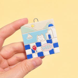 Op de afbeelding: Een vierkant keramisch ornament met een mediterrane kustscène met een witte kat, een zeilboot, een gele zon en een gebouw met een blauwe koepel. De voorgrond toont een blauw-wit geblokt patroon met een rood wijnglas en een vis.