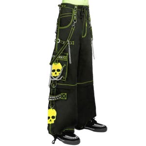 Puede incluir: Pantalones cargo negros con costuras en contraste verde neón, cadenas de metal plateado y varias correas. Los pantalones muestran gráficos de calaveras amarillas en los bolsillos laterales y la parte inferior, con zapatillas negras.