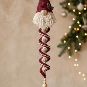 May include: A hanging crochet ornament featuring a gnome with a maroon pointed hat, a cream-colored yarn beard, and a round nose. Below the gnome is a maroon and cream spiral tail, wooden beads, and a white crocheted snowflake at the bottom.
