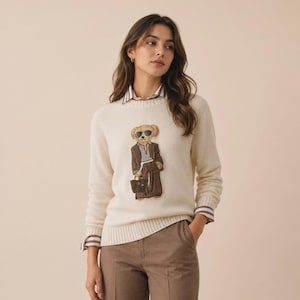 Könnte beinhalten: Ein cremefarbener Langarmpullover mit Rundhalsausschnitt und geripptem Saum. Der Pullover zeigt eine zentrale Stickerei eines Teddybären, der eine Sonnenbrille und einen braunen Anzug trägt und eine kleine braune Handtasche in der Hand hält.