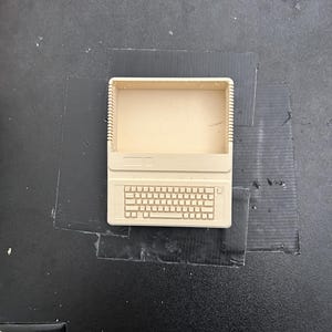 May include: A beige, rectangular, hollow container shaped like a vintage computer monitor with an integrated keyboard design on the front surface. The object features a screen-like opening at the top and a detailed grid representing keys below.