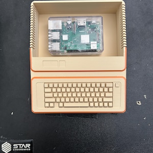 Peut inclure: Un boîtier d'ordinateur beige imprimé en 3D avec une bordure orange, doté d'un clavier intégré et d'un compartiment supérieur ouvert contenant une carte Raspberry Pi dans un boîtier de protection en plastique transparent, vu de dessus.