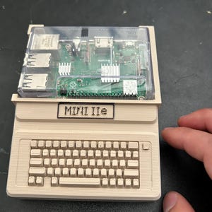 May include: A beige 3D-printed computer case shaped like a vintage desktop, featuring a built-in keyboard and a clear plastic top cover. A green Raspberry Pi circuit board is visible inside the case. The front panel displays the text MINI IIe.