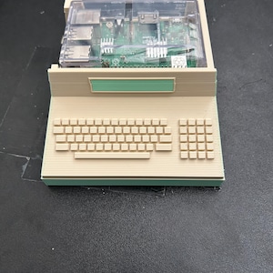 Może przedstawiać: Beżowa obudowa komputerowa wydrukowana w 3D, mieszcząca płytkę Raspberry Pi pod przezroczystą plastikową pokrywą. Podstawa posiada jasnozielony pasek akcentujący oraz formowaną klawiaturę z blokiem numerycznym i prostokątny jasnozielony panel.