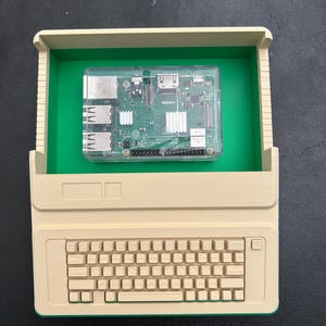 Może przedstawiać: Beżowa obudowa w kształcie komputera wydrukowana w technologii 3D z zielonym wnętrzem, wyposażona w zintegrowaną klawiaturę oraz przezroczystą plastikową osłonę zawierającą zieloną płytkę Raspberry Pi z widocznymi portami i radiatorami.