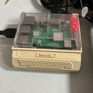 May include: A Raspberry Pi single-board computer housed in a clear plastic case, mounted on a beige 3D-printed base shaped like a vintage computer with a keyboard design. The circuit board is green with silver heat sinks and a glowing red LED light.