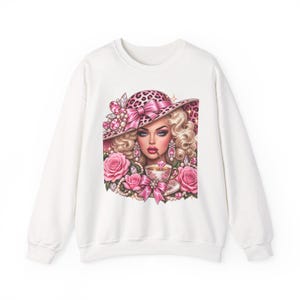 Peut inclure: Un sweat-shirt blanc à col rond orné d'une impression graphique représentant une personne aux cheveux blonds portant un chapeau à motif léopard avec un nœud rose. Le motif inclut des roses roses, des perles, des gemmes et une tasse à thé.