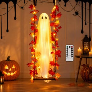 Peut inclure: Une figurine de fantôme blanche drapée de tissu, éclairée de l'intérieur, est assise sur une petite balançoire en bois suspendue par des cordes ornées de feuilles d'érable orange et rouges et de guirlandes lumineuses. Une télécommande est visible.