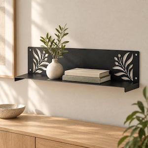 May include: A matte black metal wall shelf featuring a horizontal design with laser-cut leaf patterns on both ends. The shelf holds a small, round, off-white ceramic vase containing green foliage and two stacked books with light-colored covers.