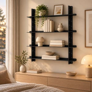 May include: A black wall-mounted shelving unit with four horizontal shelves supported by two vertical side rails. The shelves hold a trailing green plant, a framed print, books, a ceramic vase, a ribbed lamp, and a small bowl.