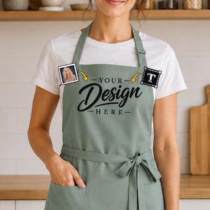 Könnte beinhalten: Eine salbeigrüne Schürze mit Taillenband, einer Vordertasche und dem schwarzen Aufdruck "YOUR DESIGN HERE" auf der Brust. Die Schürze wird über einem weißen T-Shirt getragen und bietet eine anpassbare Fläche für persönliche Grafiken und Text.