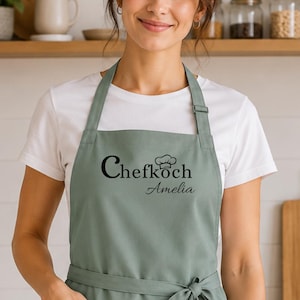 Könnte beinhalten: Eine salbeigrüne Schürze über einem weißen T-Shirt. Die Schürze hat eine Vordertasche, ein Taillenband und den schwarzen Schriftzug Chefkoch mit einem kleinen Kochmützen-Symbol über dem Namen Amelia. Das Design ist mittig auf der Brust platziert.