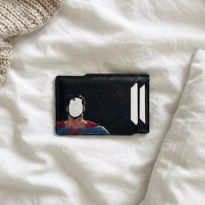 May include: A black rectangular card holder featuring a painted graphic of a figure in a red and blue suit with a blank white face. The design includes two vertical white stripes on the right side of the black leather-like surface.