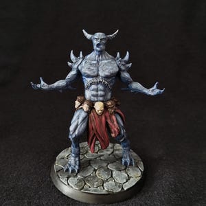 May include: A painted miniature figurine of a blue-skinned horned demon with a muscular build, standing on a stone-textured circular base. The figure wears a red loincloth adorned with a belt of small skulls and has spikes on its shoulders.