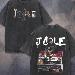 May include: A charcoal gray short-sleeve t-shirt made of 100% cotton. The front features the text J. COLE with graphic letters and a signature. The back displays the text J. COLE in white dripping font above a graphic of a person sitting on books.