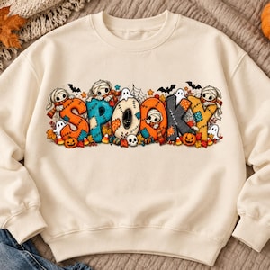 May include: A cream-colored crewneck sweatshirt featuring a large, colorful graphic across the chest. The graphic displays the word SPOOKY in patchwork-style letters, decorated with illustrations of mummies, ghosts, bats, pumpkins, and autumn leaves.