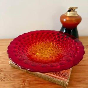 May include: A round glass bowl featuring a vibrant red and orange ombre colour gradient with a raised hobnail texture across the surface. The bowl has a scalloped edge and sits atop a rectangular book with a patterned cover and gold accents.