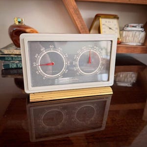 May include: A rectangular Airguide weather station with a white frame and a gold-toned base. The device features two circular gauges on a dark grey background, one labelled Humidity and the other Temperature, each with a red needle indicator.