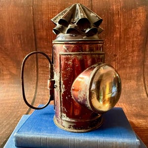 Peut inclure: Une lanterne en métal vintage avec une finition rouge foncé et des signes d'usure visibles. Elle présente une lentille en verre circulaire proéminente sur le devant, une grande poignée latérale incurvée et un capuchon supérieur en métal.