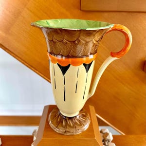 Peut inclure: Un pichet en céramique avec une base crème ornée de motifs de gouttes verticales noires, une bande orange et un bord festonné marron. L'intérieur est vert clair et l'anse est orange. La base présente un motif festonné marron texturé.