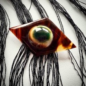 May include: A diamond-shaped brooch featuring a glossy, tortoiseshell-patterned brown resin base with a central, raised, spherical eye-like element. The eye has a dark green iris, a white sclera, and a black pupil, set against black fringe.