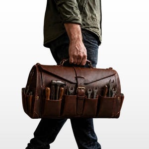May include: A dark brown leather tool bag featuring a top handle, a central buckle strap, and multiple exterior pockets. The pockets contain various metal hand tools, including a hammer, several wrenches, and wooden-handled screwdrivers.
