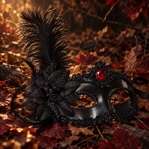 Pode incluir: Uma máscara de baile de máscaras preta com uma pluma alta preta, um grupo de flores de tecido preto e contas pretas intrincadas ao longo das bordas. Uma única pedra preciosa vermelha de formato oval está colocada no centro da testa.