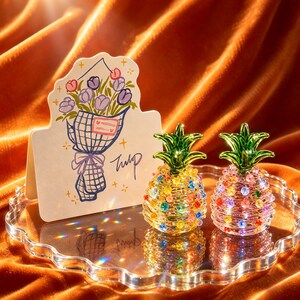 Könnte beinhalten: Zwei Ananas-Figuren aus Glas mit grünen Spitzen und bunten eingebetteten Kristallen, neben einer weißen Grußkarte mit einer handgezeichneten Illustration eines Tulpenstraußes und dem handschriftlichen Text Tulip, auf einem gewellten Tablett.