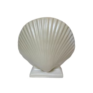 May include: A cream-colored ceramic decorative object shaped like a large scallop shell. The piece features a glossy finish with prominent, radiating vertical ridges that extend from the base to the curved top edge, supported by a square pedestal.